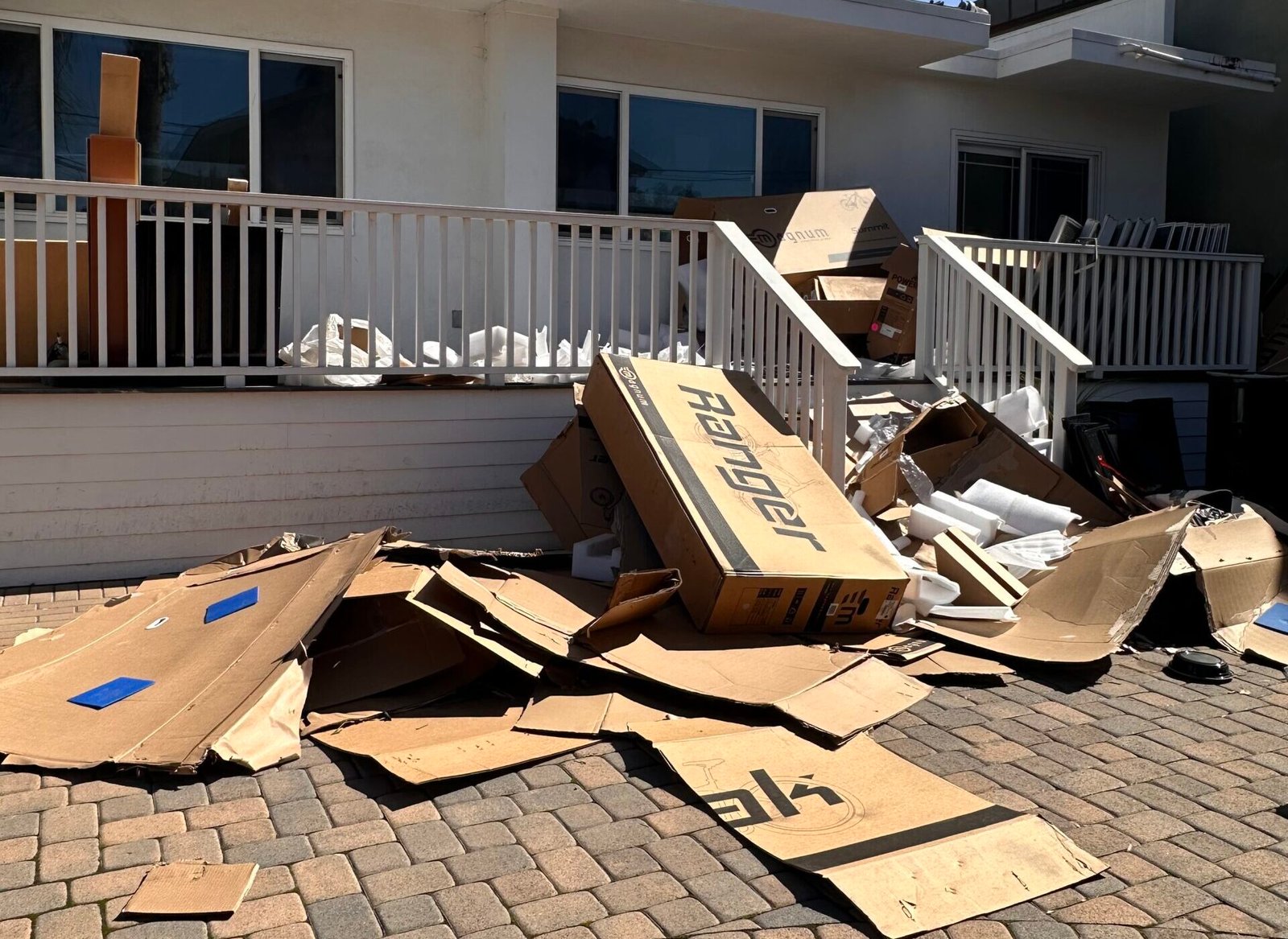 A residential front yard with extensive cardboard debris and packing materials piled on a paved driveway in front of a white house with a railing.