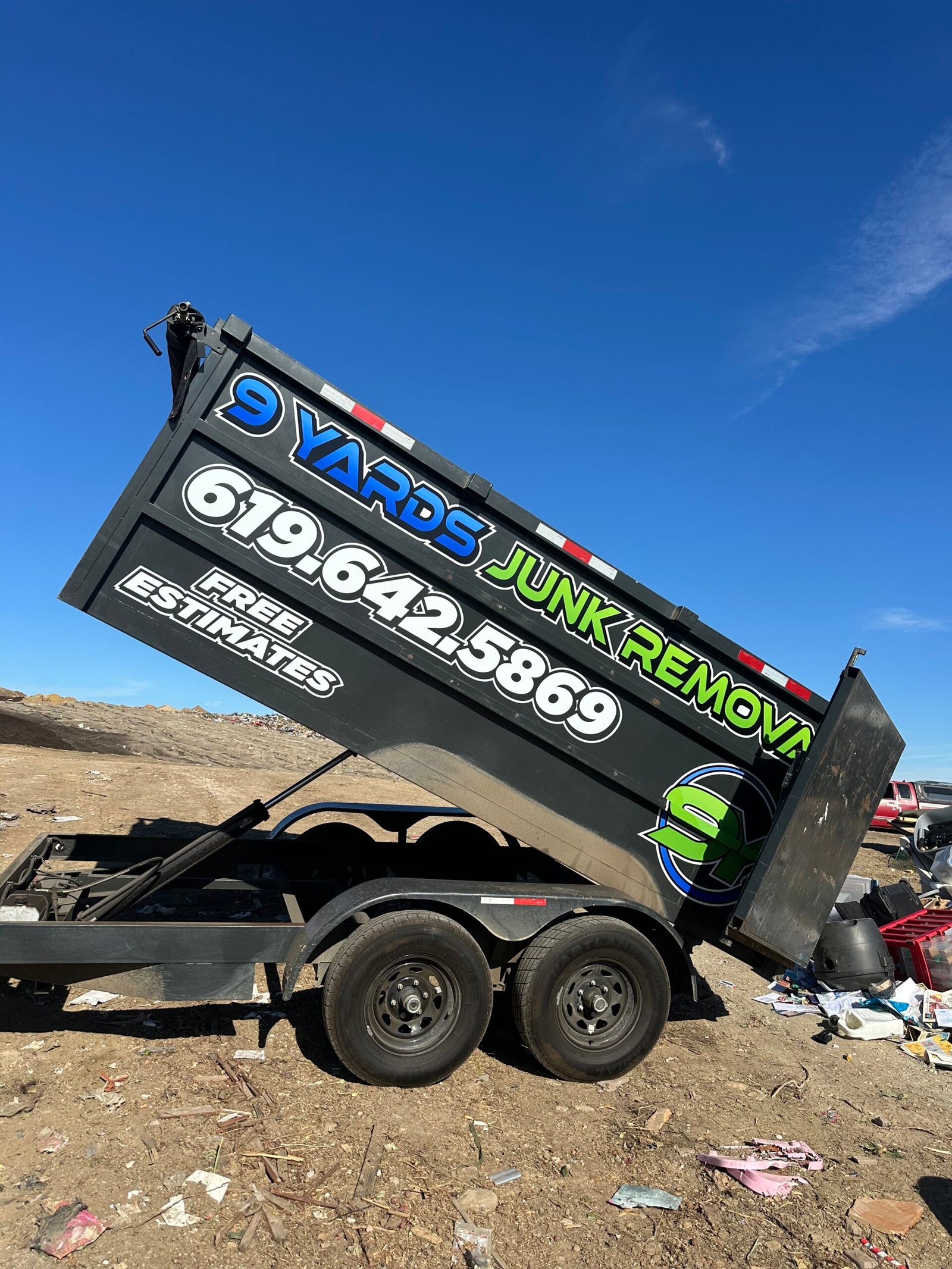 A dark green trailer and pickup truck parked in a parking lot, features branding for a junk removal service. The trailer has bold neon green and blue lettering reading “Junk Removal” with a large circular logo and the phone number 619-642-5869.
