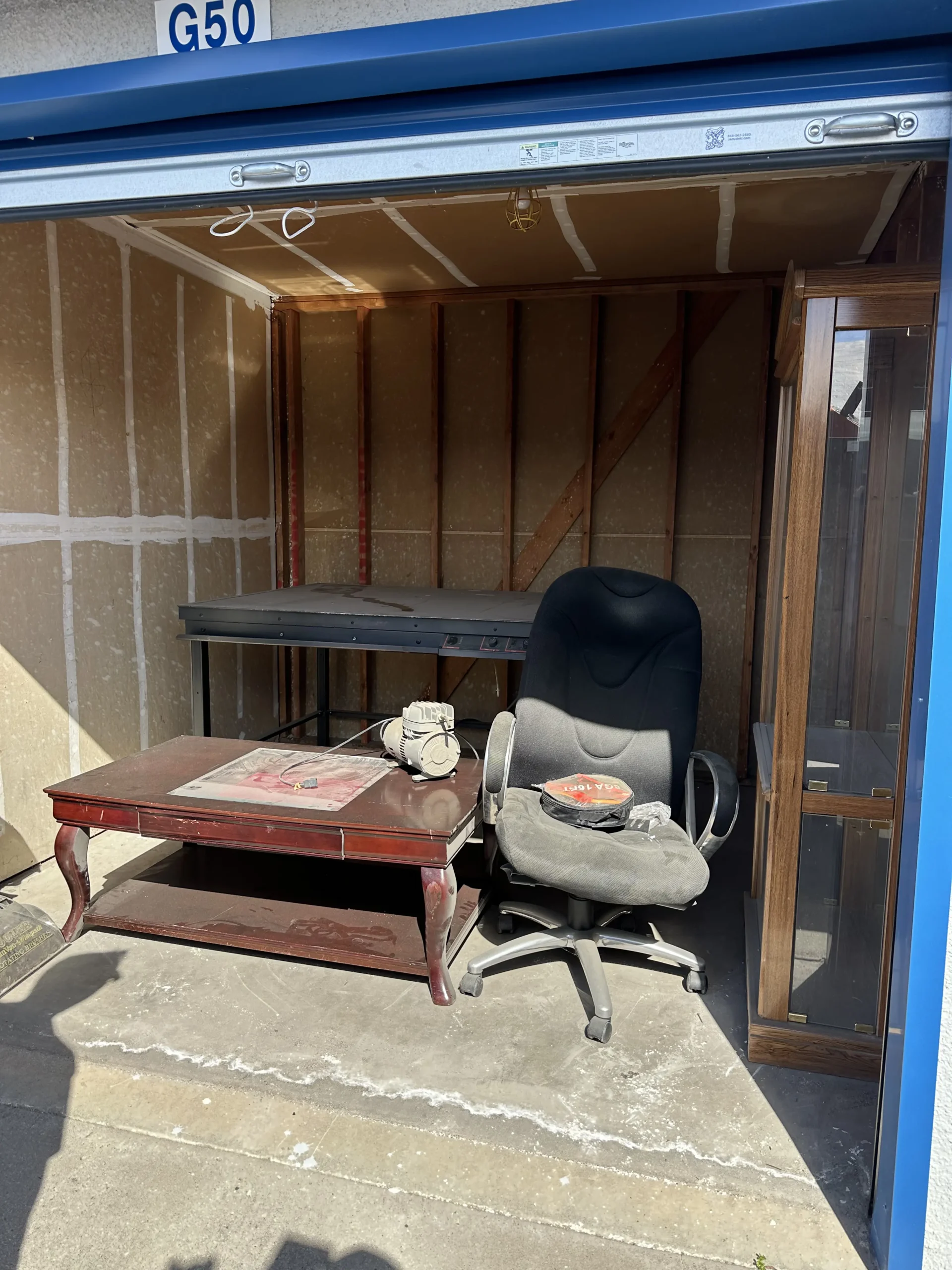 Large Appliances Junk Removal A small storage unit with a blue frame labeled “G50” above the doorway. Inside, cluttered with office furniture including a worn black swivel chair, a red wooden coffee table with papers on top, a metal workbench along the back wall, and a wooden cabinet leaning against the right wall. The interior walls show exposed studs and diagonal bracing. The unit is outdoors with sunlight casting shadows on the concrete floor.