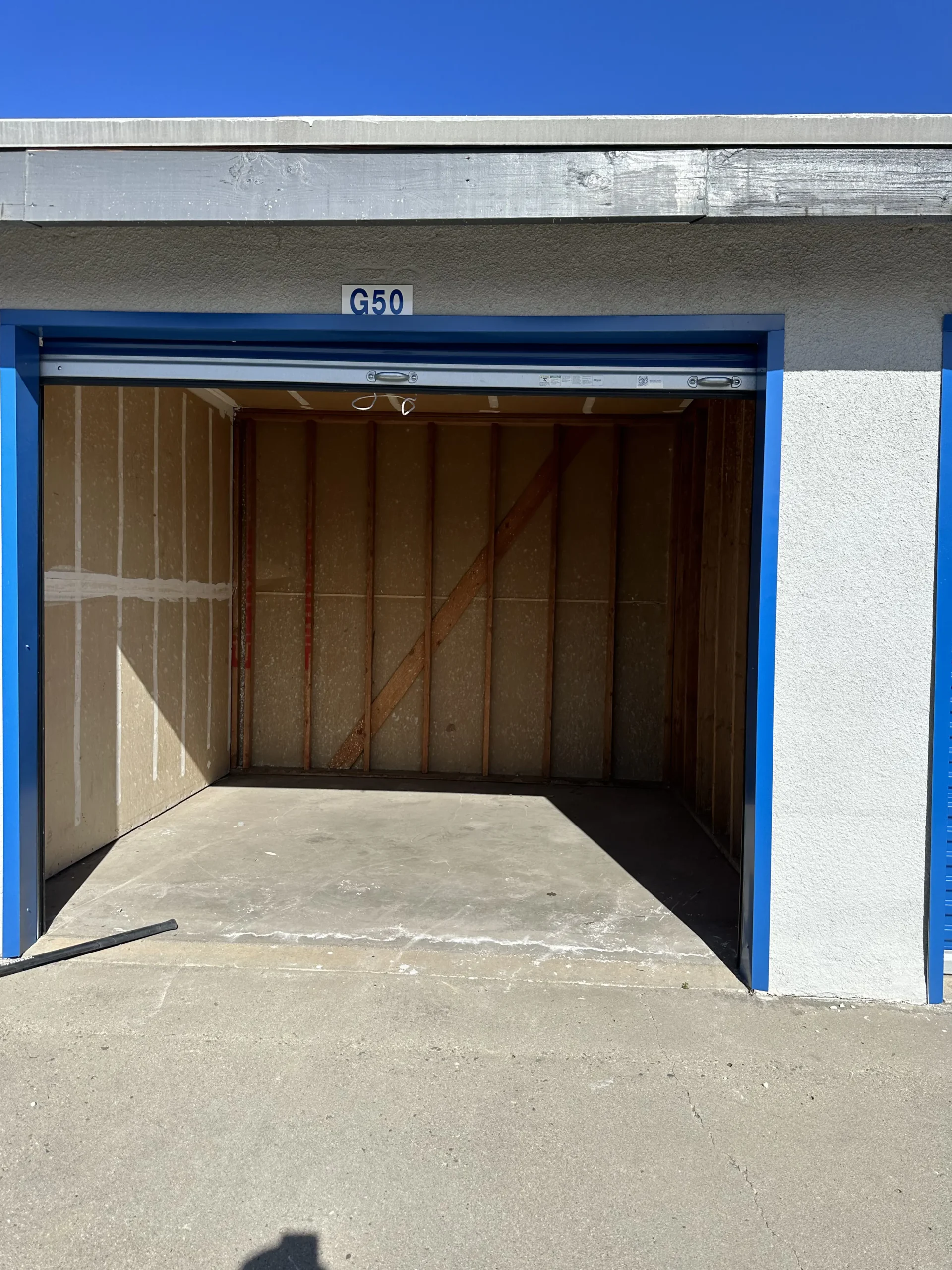 Large Appliances Junk Removal An empty self-storage unit with a blue frame and white walls. The roll-up door is open, revealing a bare interior with wooden studs and diagonal bracing. The unit is labeled “G50” above the doorway. The surrounding exterior wall is light-colored with blue trim, and the ground in front is a concrete surface under a bright sky.