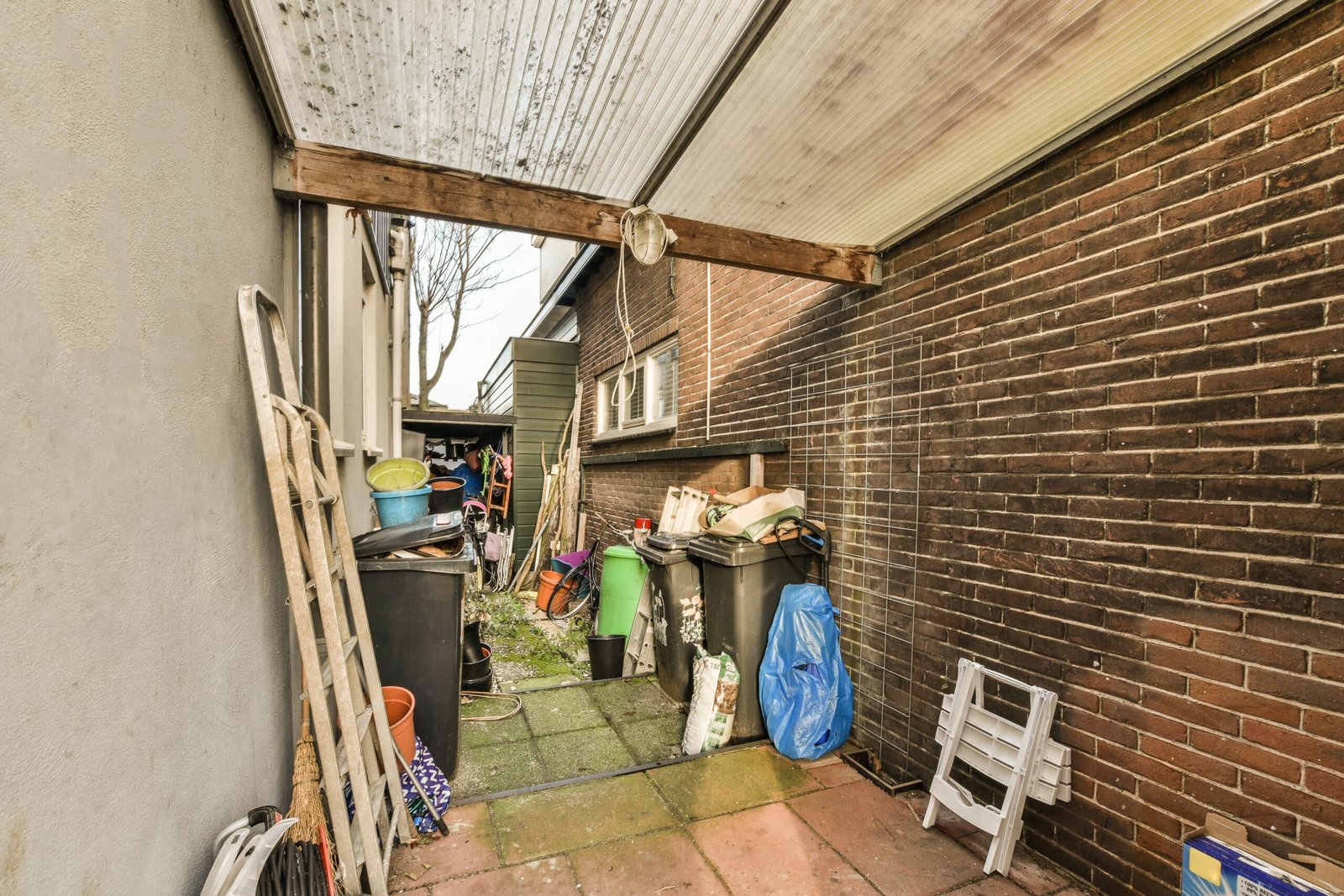 Home Cleanouts A narrow outdoor storage area/range between two brick walls, cluttered with assorted junk. Left side shows a wooden ladder leaning against the wall, buckets, and various tools. Center and right sections are filled with trash cans, bags, boxes, and scattered debris.