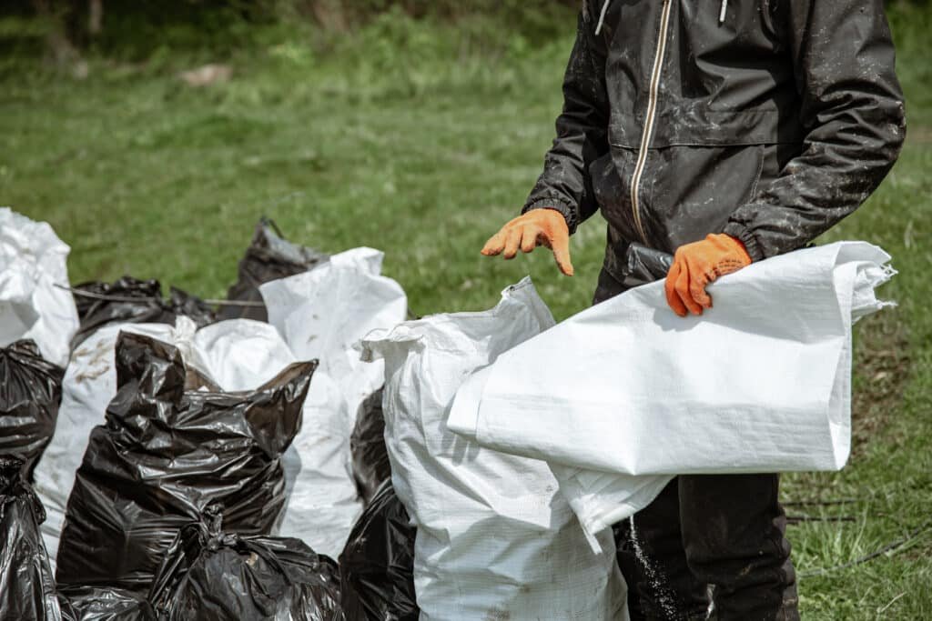 What Items Can Junk Removal Companies Take? A person wearing a dark waterproof suit and orange gloves stands outdoors, folding or handling a large white plastic bag or tarp.