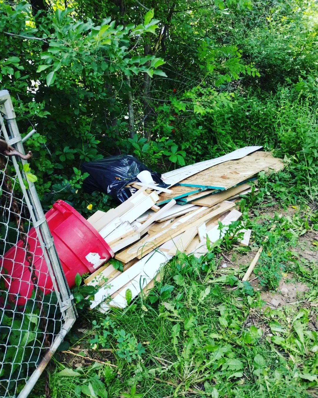 Construction Debris Removal pile of wooden boards and miscellaneous debris stacked along a chain-link fence in a grassy, overgrown outdoor area.