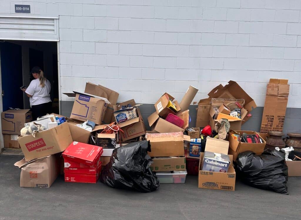 A large pile of cardboard boxes and black trash bags stacked outside a light gray building, with a doorway on the left where a person is standing inside.