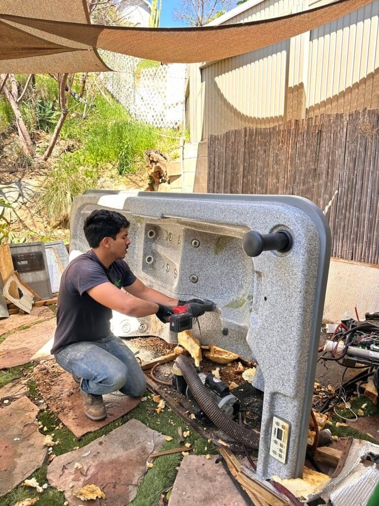 Man kneeling outdoors, repairing a large pool equipment or spa device with tools.