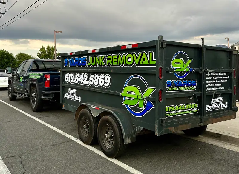 A dark green trailer and pickup truck parked in a parking lot, features branding for a junk removal service. The trailer has bold neon green and blue lettering reading “Junk Removal” with a large circular logo and the phone number 619-642-5869.
