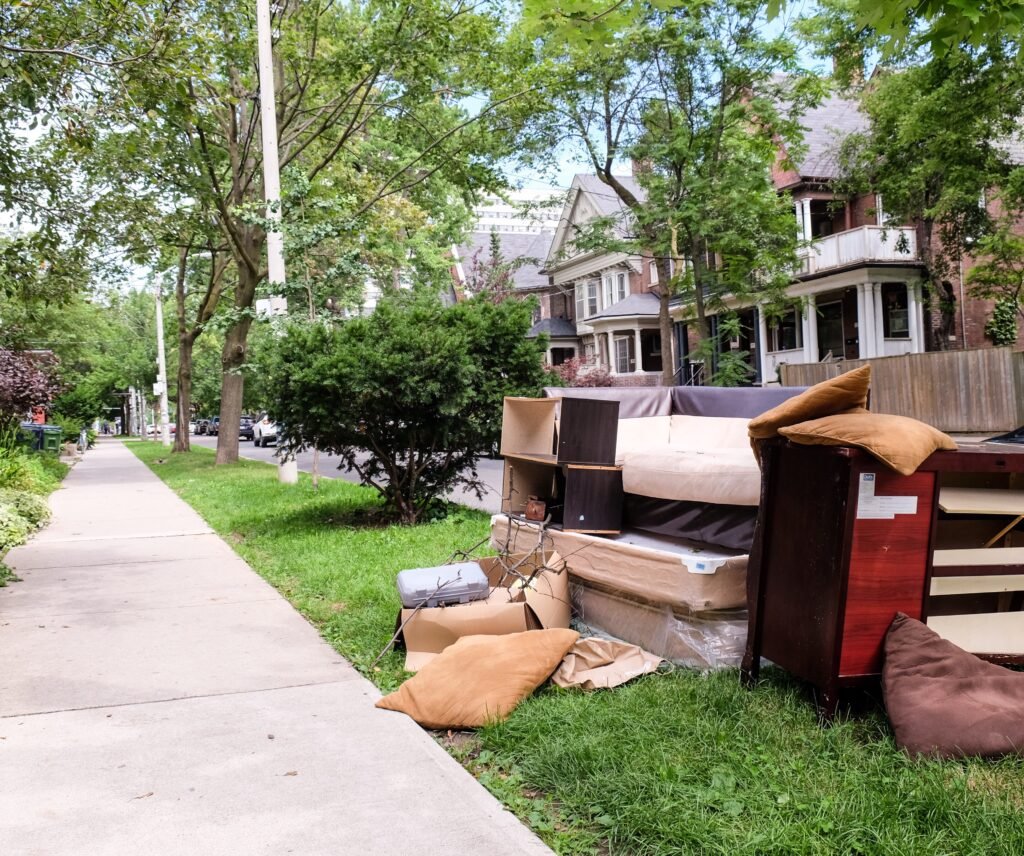 A residential sidewalk scene with a pile of discarded furniture and bags along the grassy curb.