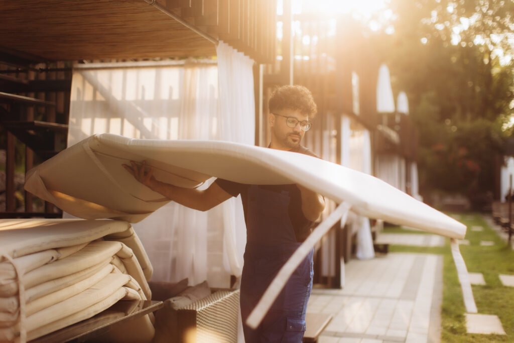 Hotel employee carrying a white mattress outdoors, positioned near flowing white curtains as the sun sets, casting warm light across the scene