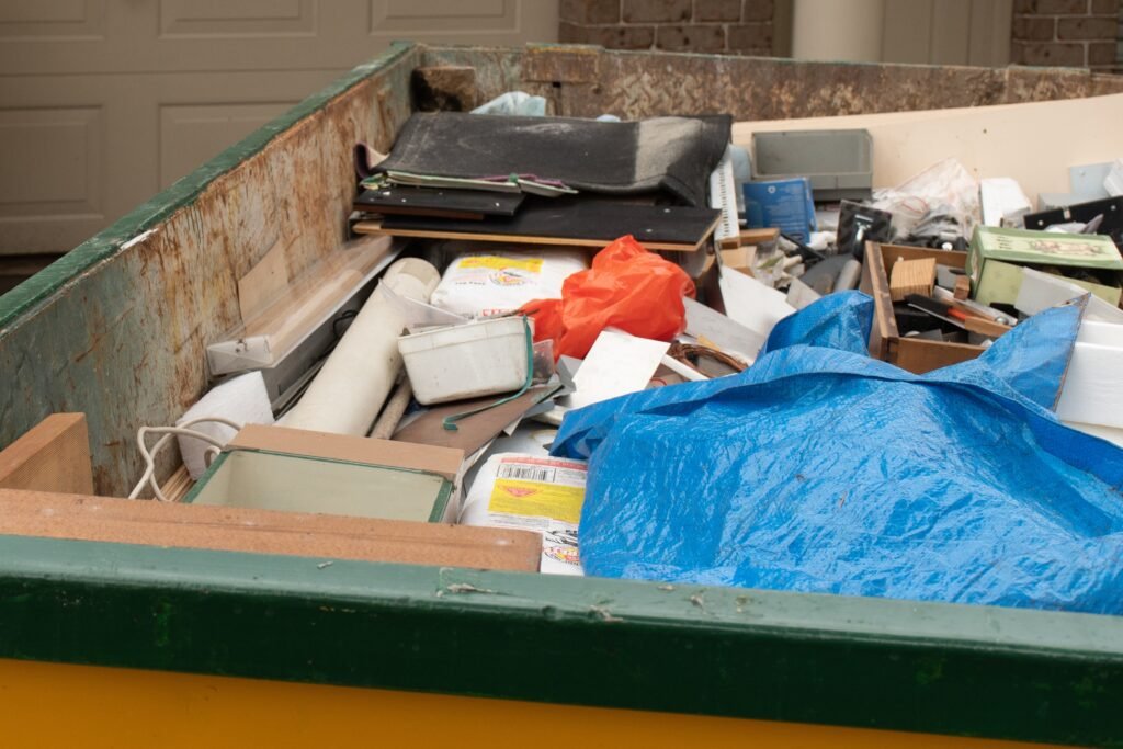 A cluttered dumpster filled with various discarded items, including foam, pieces of wood, cardboard, a blue tarp, and miscellaneous debris, with a brick wall and garage door in the background.